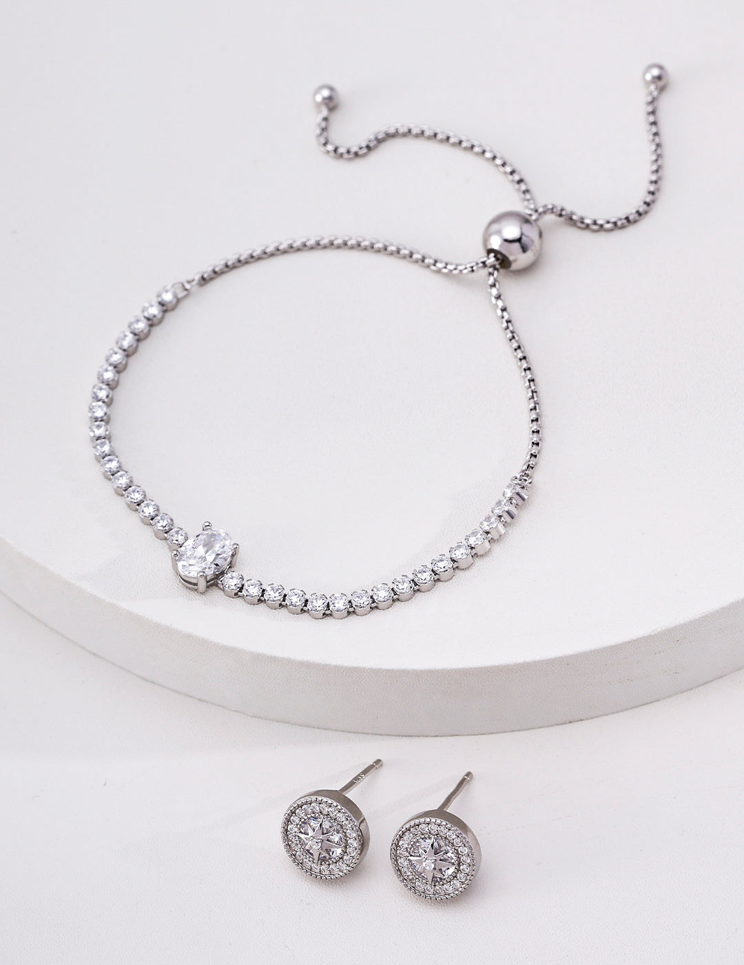 Silver cubic zirconia tennis bracelet displayed beside round halo stud earrings